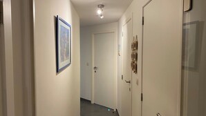 Un couloir avec un tableau encadré sur le mur de gauche, une porte au fond et un luminaire au plafond.