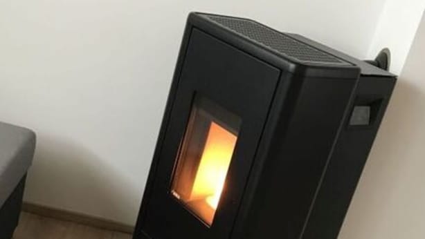 A black wood-burning stove with a visible fire inside, placed on a wooden floor next to a white wall. - Baier Holiday House (Philippsreut)