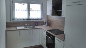 A compact kitchen with white cabinets, a built-in oven, a coffee maker, and a window with curtains. - Baier Holiday House (Philippsreut)