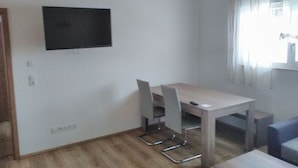 A room with a wooden ceiling, a flat-screen TV mounted on the wall, a wooden dining table with chairs, and a window with curtains. - Baier Holiday House (Philippsreut)