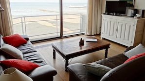 Ett vardagsrum med en stor skjutdörr i glas som leder ut till en terrass med utsikt över stranden, en soffa med röda och grå kuddar, ett soffbord i trä och en tv-apparat placerad på ett skåp.