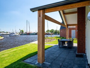 A covered outdoor area with a table and chairs, a brick building, and a view of boats on the water. - House for the Disabled, Holiday Park in Friesland (Akkrum)
