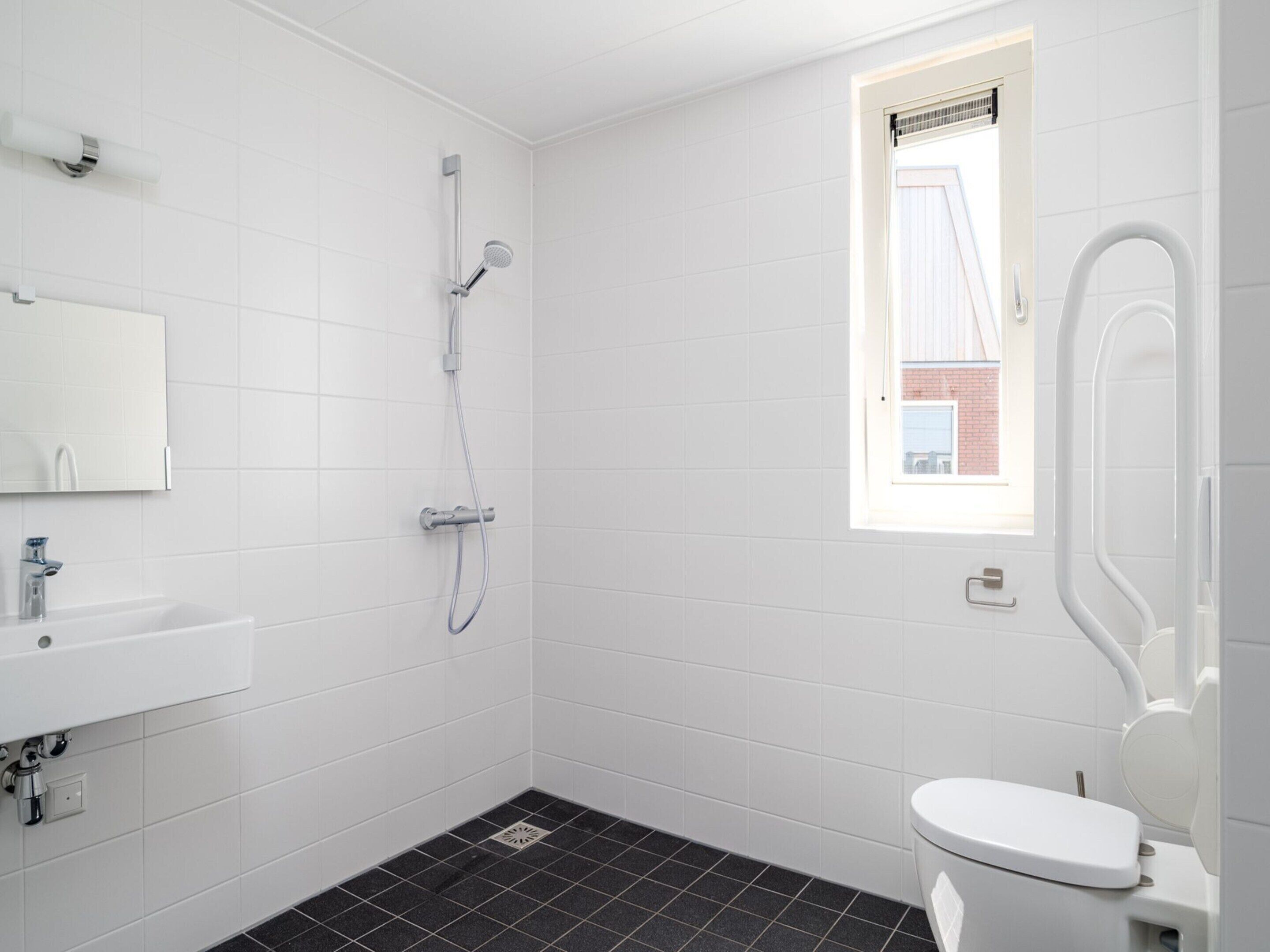 A modern bathroom with white tiles, a black floor, a shower, a toilet, and a window with a view of a building.