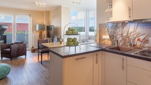 Eine moderne Küche mit Kochinsel, ein Essbereich mit Sofa und Stühlen sowie ein Ausblick ins Freie durch große Fenster.
