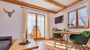 A spacious room with wooden floors, a dining table, two green chairs, a sofa, a TV mounted on the wall, and a balcony with curtains. - Holiday Apartment "wetterstein" (Kruen)