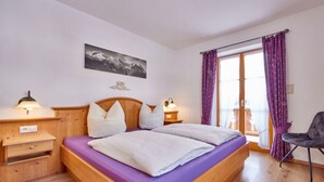 A bedroom with a wooden bed, purple bedding, a nightstand with a lamp, a chair, and a window with purple curtains. - Holiday Apartment "wetterstein" (Kruen)