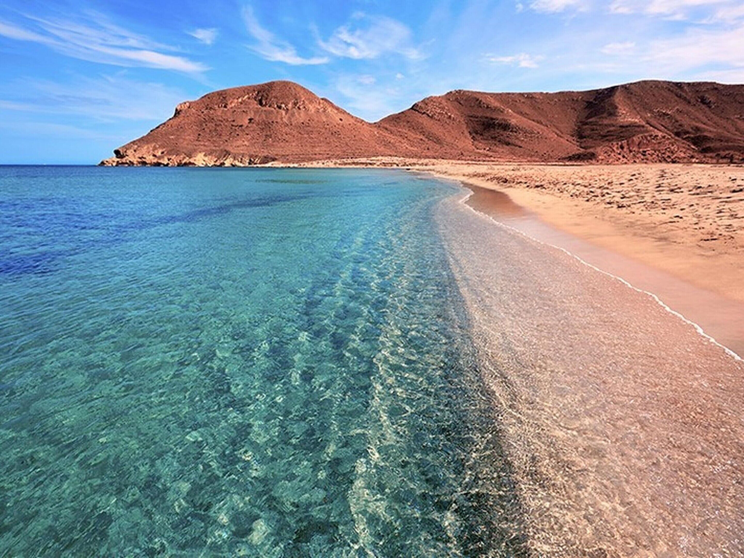 Una playa con aguas turquesas cristalinas, orilla de arena y colinas rocosas al fondo.