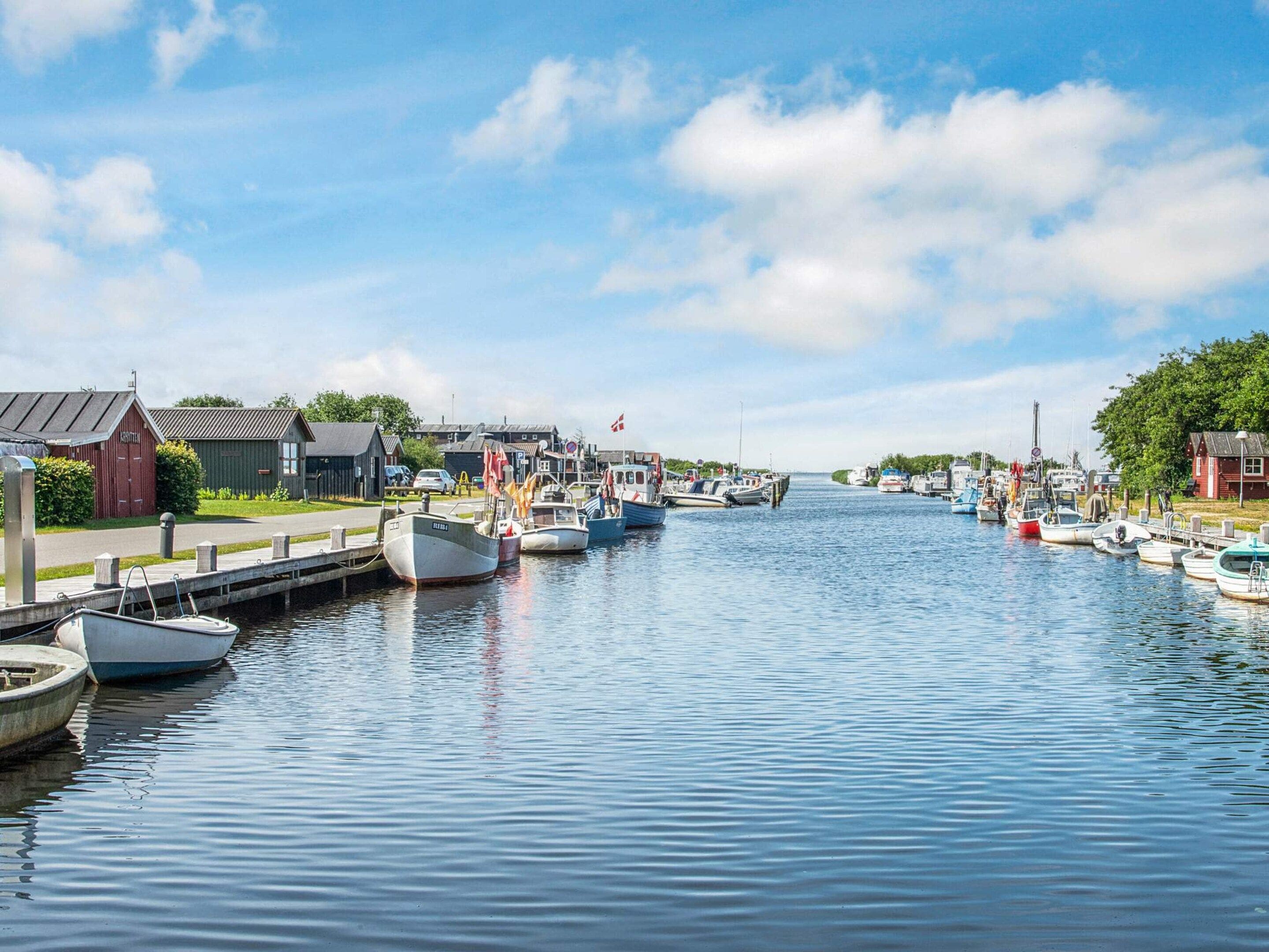 A calm waterway with boats docked on both sides, a clear sky, and buildings along the shore.