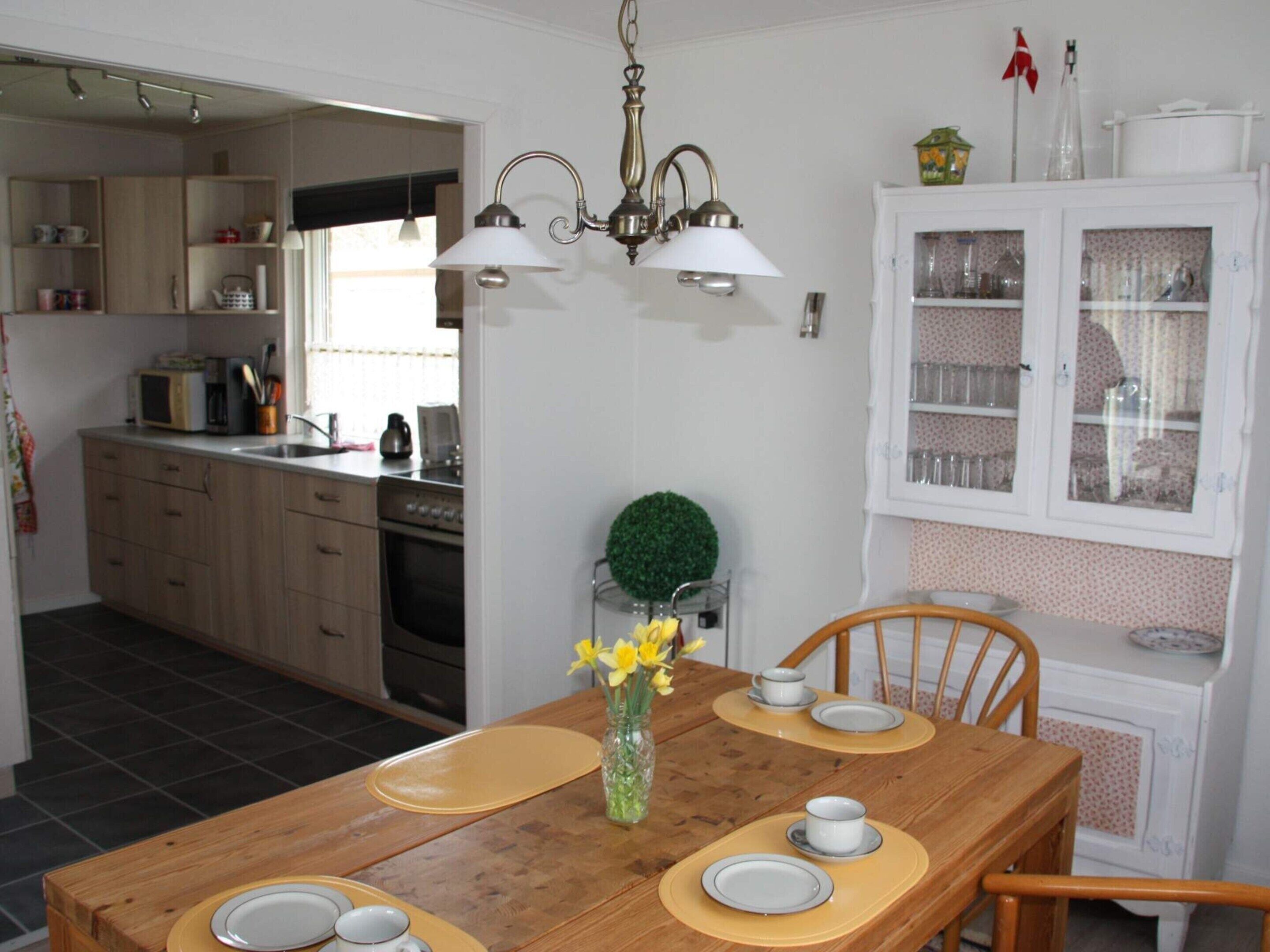 Comedor con una mesa de madera dispuesta para cuatro personas, un jarrón con flores y una cocina al fondo.
