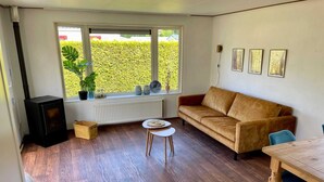 A cozy living room with a sofa, a small table, a wooden floor, a window with a view of greenery, and a black heater.