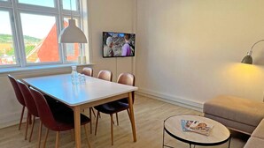 Une pièce comprenant une table à manger, des chaises, un canapé, une petite table ronde avec un magazine, un téléviseur fixé au mur et une fenêtre donnant sur des immeubles.