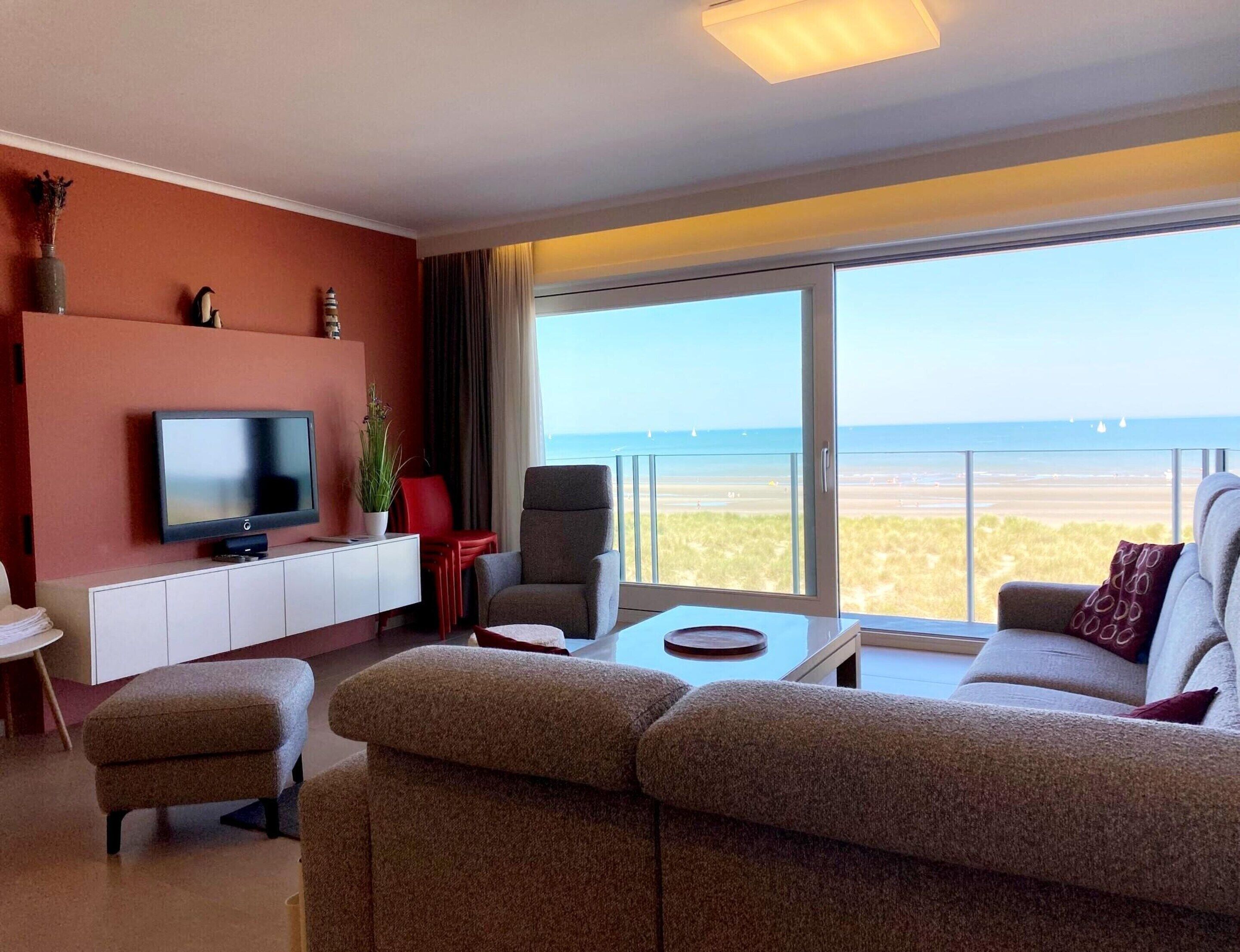 Un salon moderne avec un téléviseur à écran plat, un canapé, un fauteuil rouge et une vue sur la plage.