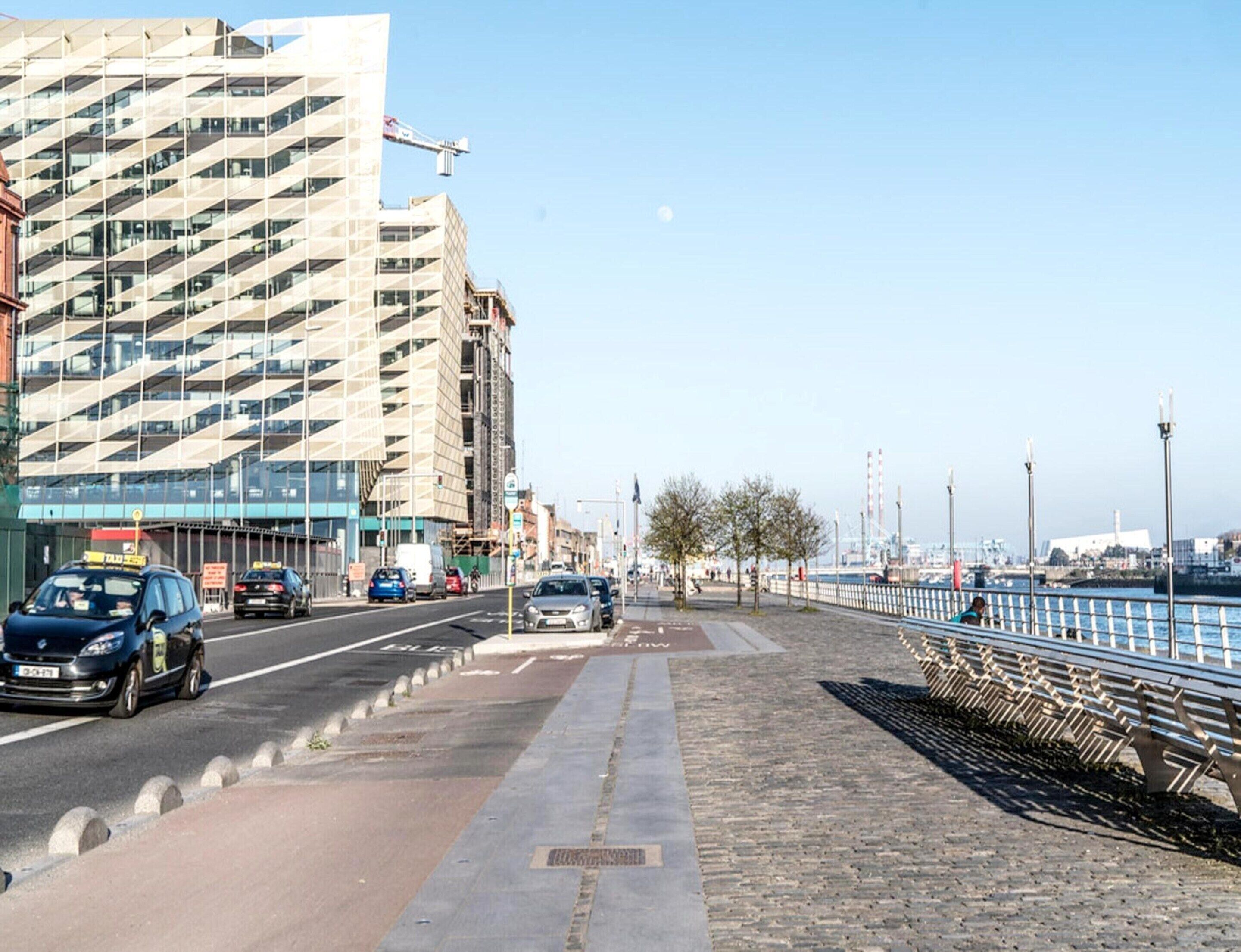 A waterfront promenade with a row of benches, a pedestrian path, and a road with cars.