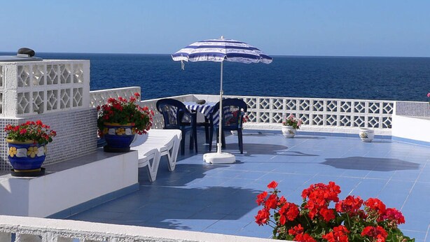 Um terraço com mesa e cadeiras, um guarda-sol azul e branco listrado, e flores vermelhas.