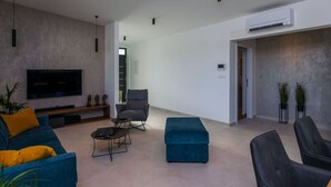 Ein modernes Wohnzimmer mit Flachbildfernseher, einem blauen Sofa, einem grauen Sessel, einem blauen Hocker und einem Couchtisch.