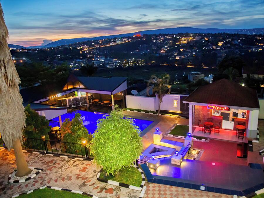 Un área de piscina en la azotea con un restaurante, rodeada de jardines ajardinados y vistas a un paisaje urbano al atardecer.