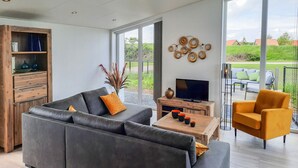 A modern living room with a grey sofa, a wooden coffee table, a TV, and a large window offering a view of the outdoors. - Detached House in Zeeland With Scenic Terrace (Wemeldinge)