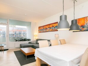 A modern hotel room with a large bed, a dining table, and a sofa. There is a painting on the wall and a window with a view of buildings outside.
