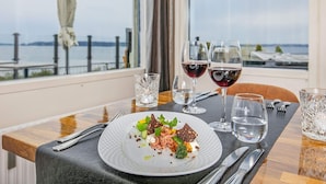 Ein Esstisch mit einem Teller Essen, zwei Gläsern Rotwein und Meerblick durch ein Fenster.