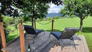 Eine Holzterrasse mit zwei Liegestühlen und einer Weinflasche, umgeben von Grün und Blick auf Hügel.