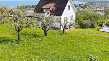 A white house with a red roof, surrounded by green grass and trees, overlooking a coastal town.