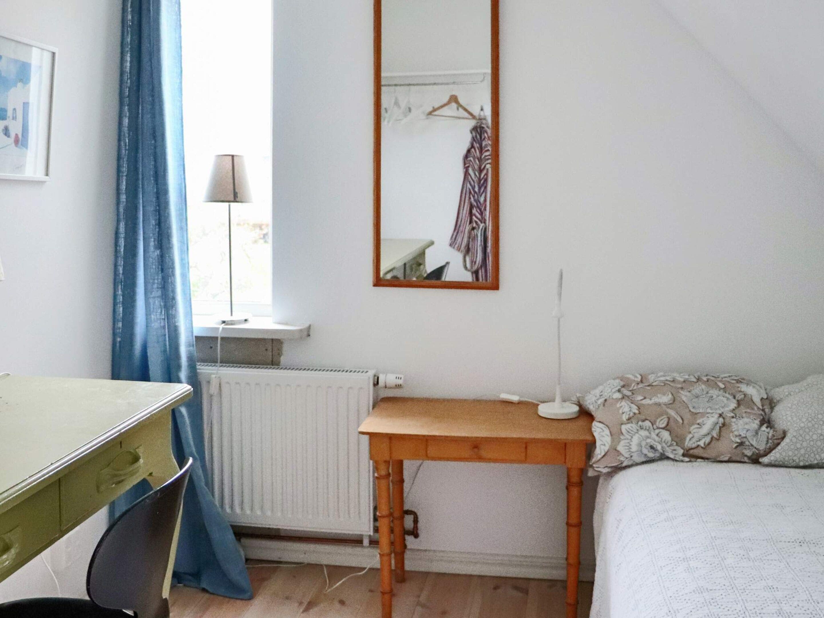 A room with a bed, a desk, a chair, a radiator, a mirror, and a window with curtains.