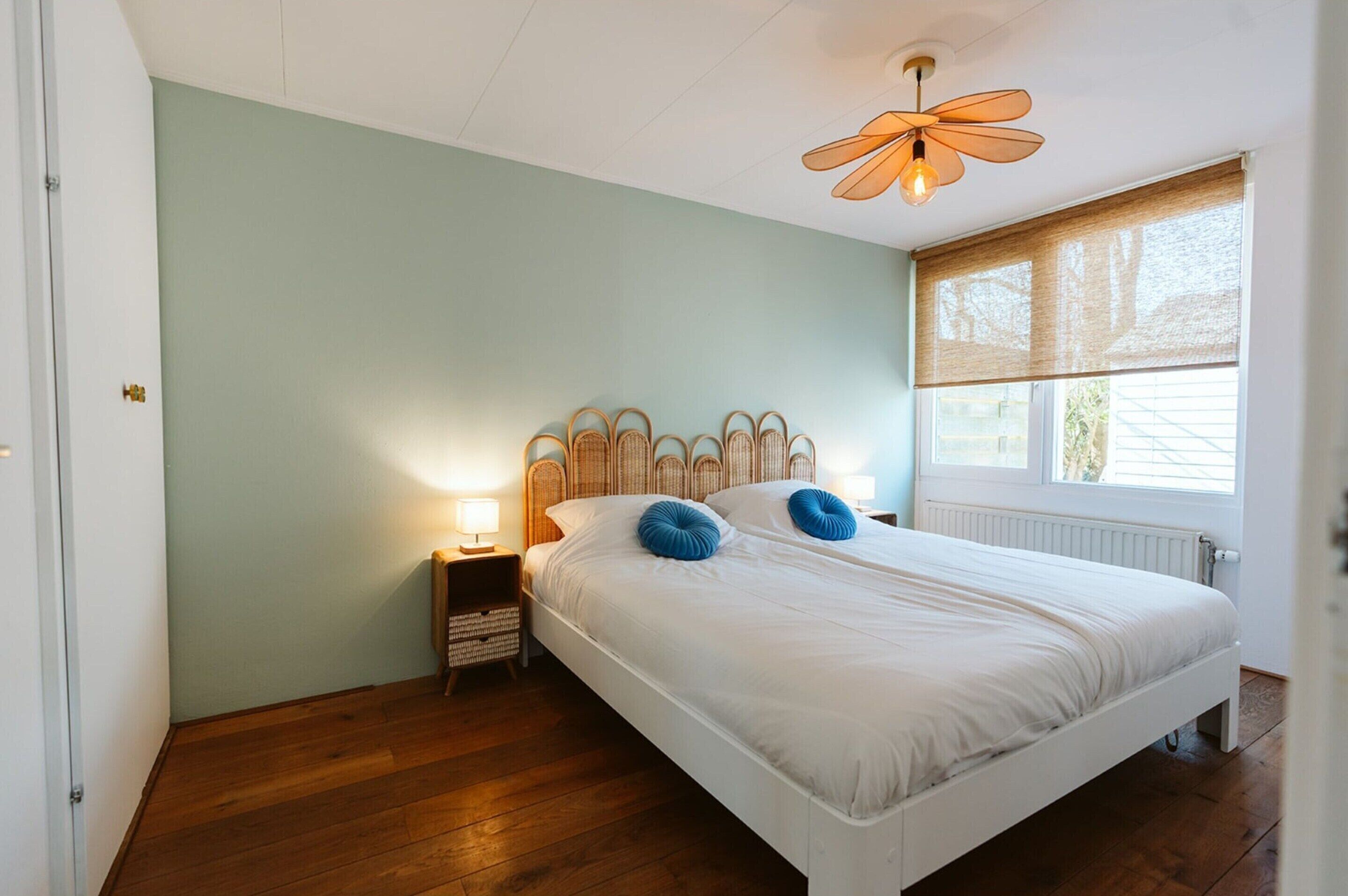 Quarto com cama branca, duas almofadas azuis, cabeceira de vime, mesa de cabeceira com abajur e janela com persianas.