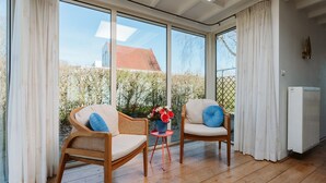 A sunlit room with two wicker chairs, a small table with a vase of flowers, and a view of a red-roofed house through large windows. - Holiday Bungalow With a Nice Light Appearance at 1.5 km From the Beach (Ouddorp)