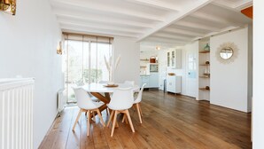 A spacious dining area with a wooden table and white chairs, a kitchen in the background, and a large window with blinds. - Holiday Bungalow With a Nice Light Appearance at 1.5 km From the Beach (Ouddorp)