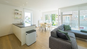 Een moderne keuken met een geïntegreerd eiland, een eethoek met een tafel en stoelen, en een zithoek met een bank en een groot raam met uitzicht op de straat.