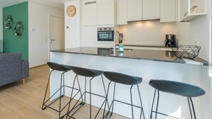 Een moderne keuken met witte kasten, een centraal eiland met barkrukken, een klok aan de muur en een koffieapparaat.