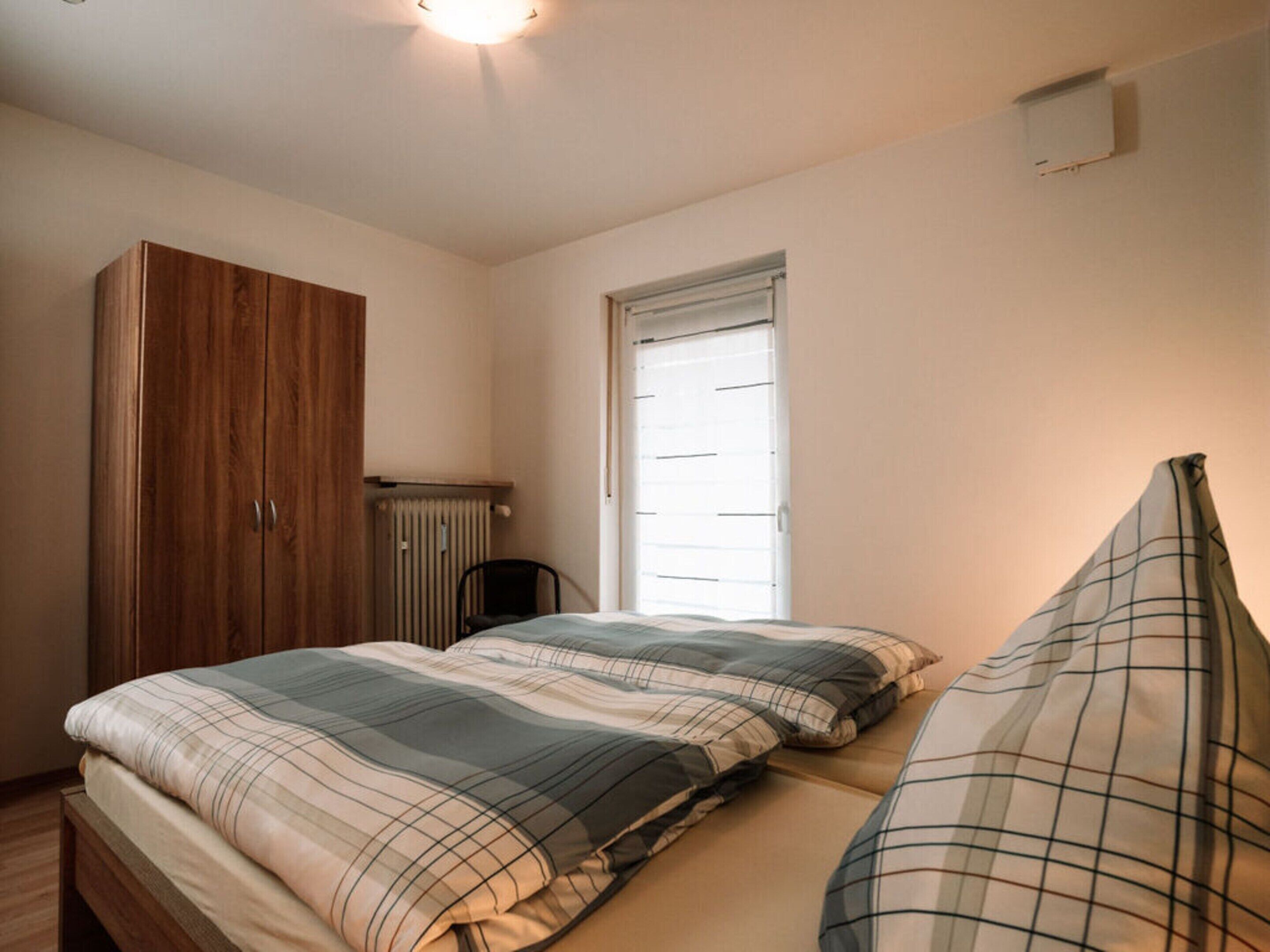 A bedroom with a wooden wardrobe, a radiator, a window, and a bed with plaid bedding.
