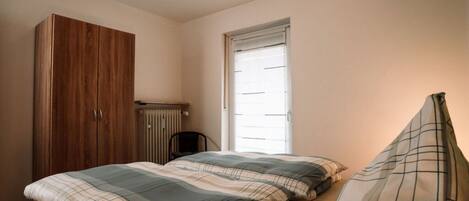 A bedroom with a wooden wardrobe, a radiator, a window, and a bed with plaid bedding.