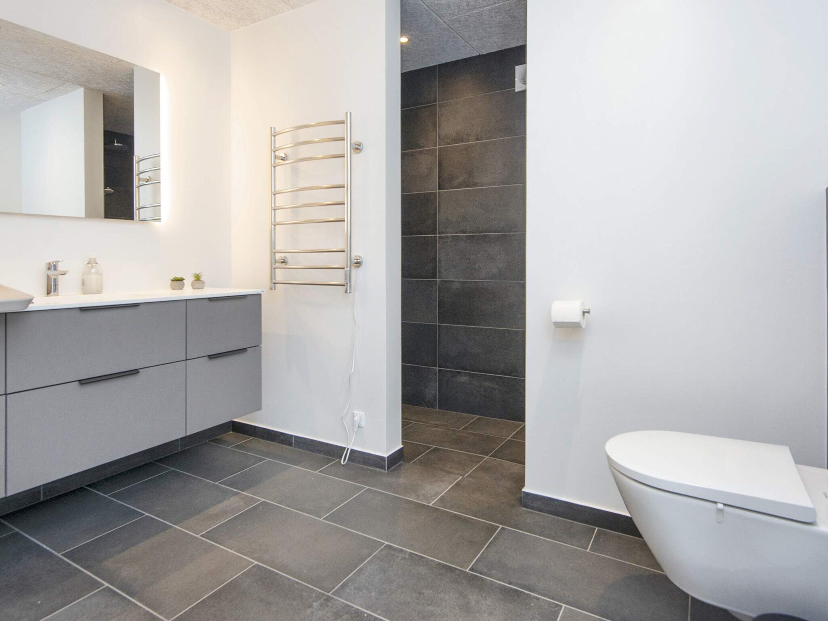 A modern bathroom with a grey tiled floor, a white toilet, a built-in sink with a mirror, and a towel rack.