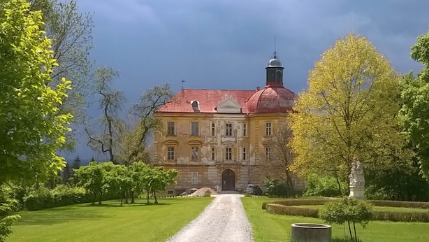 A large, historic building with a red roof and a central dome, surrounded by well-maintained lawns and trees.