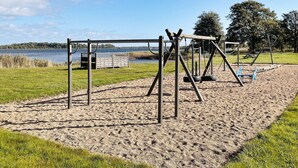 A playground with swings and a slide, set on a sandy area with grass and trees in the background. - 4 Star Holiday Home in Sturko (Sturkö)