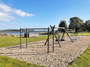 Un parque infantil con columpios y un tobogán, situado en una zona arenosa con césped y árboles al fondo.