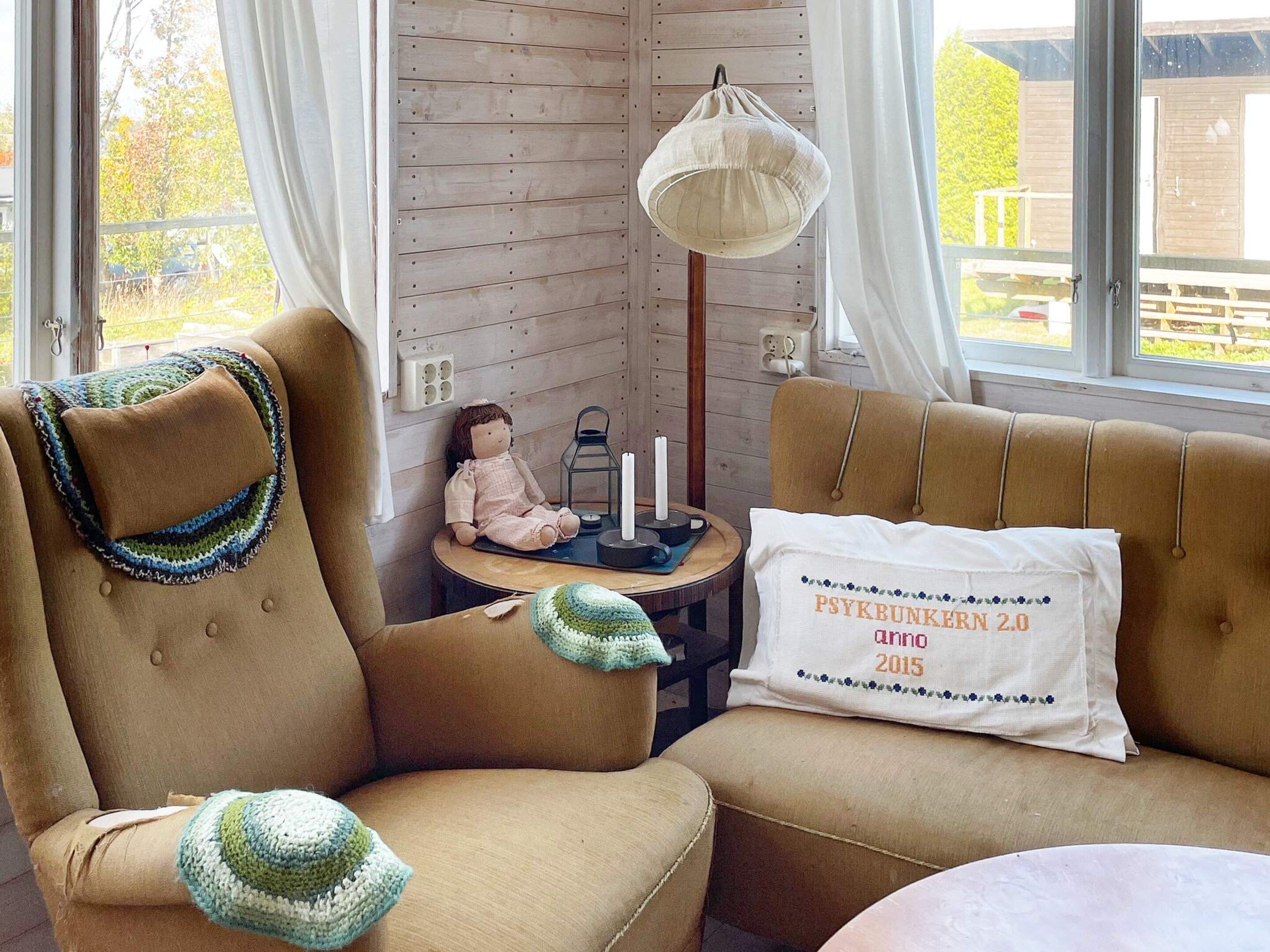 A cozy room with two brown upholstered chairs, a small table with a lamp, a candle, and a doll. There's a pillow with text on it.