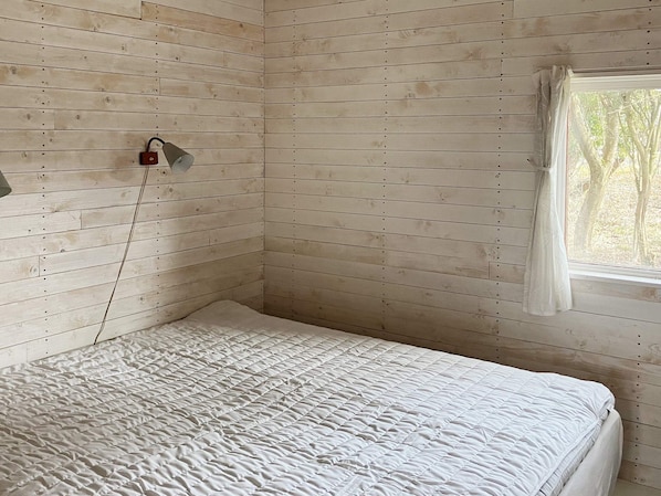 A room with a wooden slat wall, a bed with a white quilt, a window with a white curtain, and a wall-mounted light fixture. - 4 Star Holiday Home in Sturko (Sturkö)