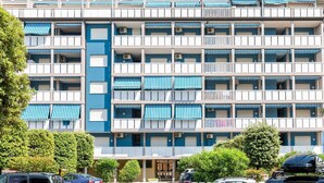 A multi-story hotel with balconies, air conditioning units, and a parking lot with several cars.