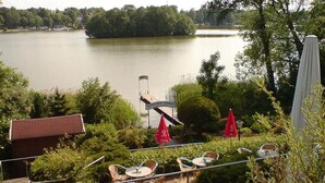 A terrace with outdoor seating overlooking a lake, surrounded by greenery and a small building with a red roof. - Amazing Apartment in Templin (Templin)