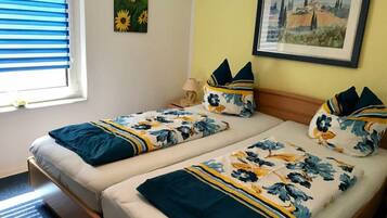 A room with two beds, each with floral bedding and matching pillows. There is a window with blinds, a painting on the wall, and a lamp on the nightstand.