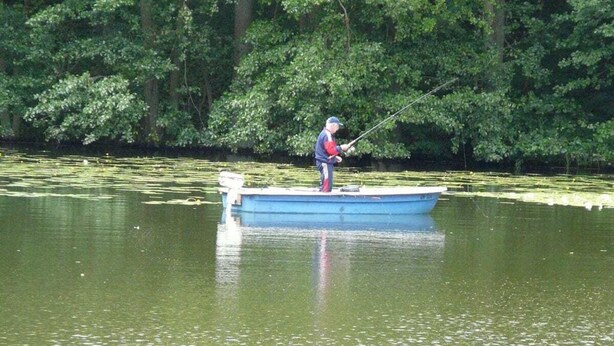 Une personne pêchant depuis un petit bateau bleu sur un lac paisible, entouré d’une végétation luxuriante.