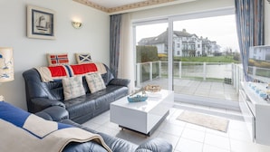 A modern living room with a leather sofa, a white coffee table, and a view of a large house across a body of water. - Cozy Apartment With Sunny Terrace (Middelkerke)