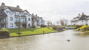A canal with a row of modern houses on one side and a grassy area with a small tree. - Cozy Apartment With Sunny Terrace (Middelkerke)