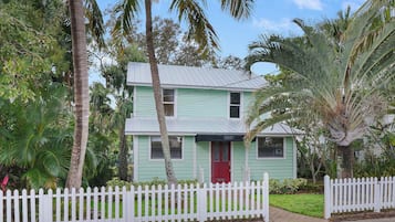 A two-story house with a red front door, surrounded by a white picket fence, and palm trees in the background.