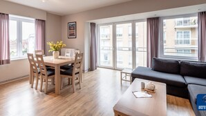 A modern apartment with a dining area, a sofa, and large windows offering a view of the city. - Goethe 0202 Furnished Apartment With sea View (De Haan)