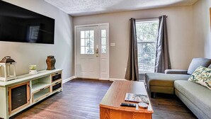 Un salon avec un téléviseur à écran plat fixé au mur, une table basse en bois, un canapé et une fenêtre avec des rideaux.