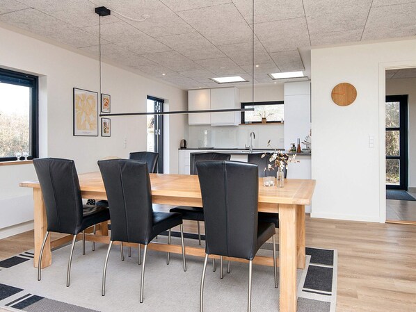 Un espace repas moderne avec une table en bois et des chaises noires, une cuisine en arrière-plan et une horloge au mur.