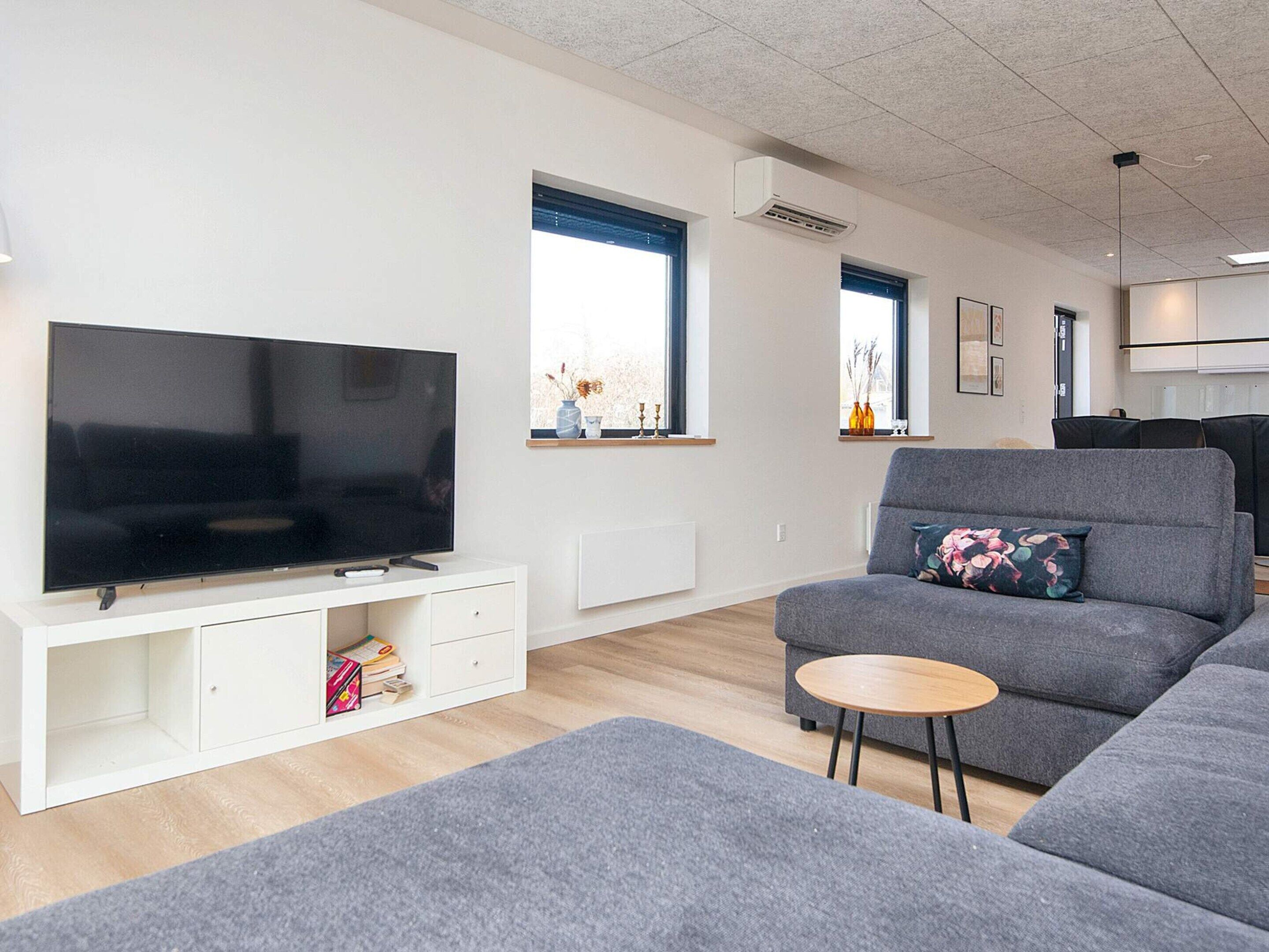 A modern living room with a flat-screen TV on a white cabinet, a grey sofa with a floral pillow, and a small wooden side table.